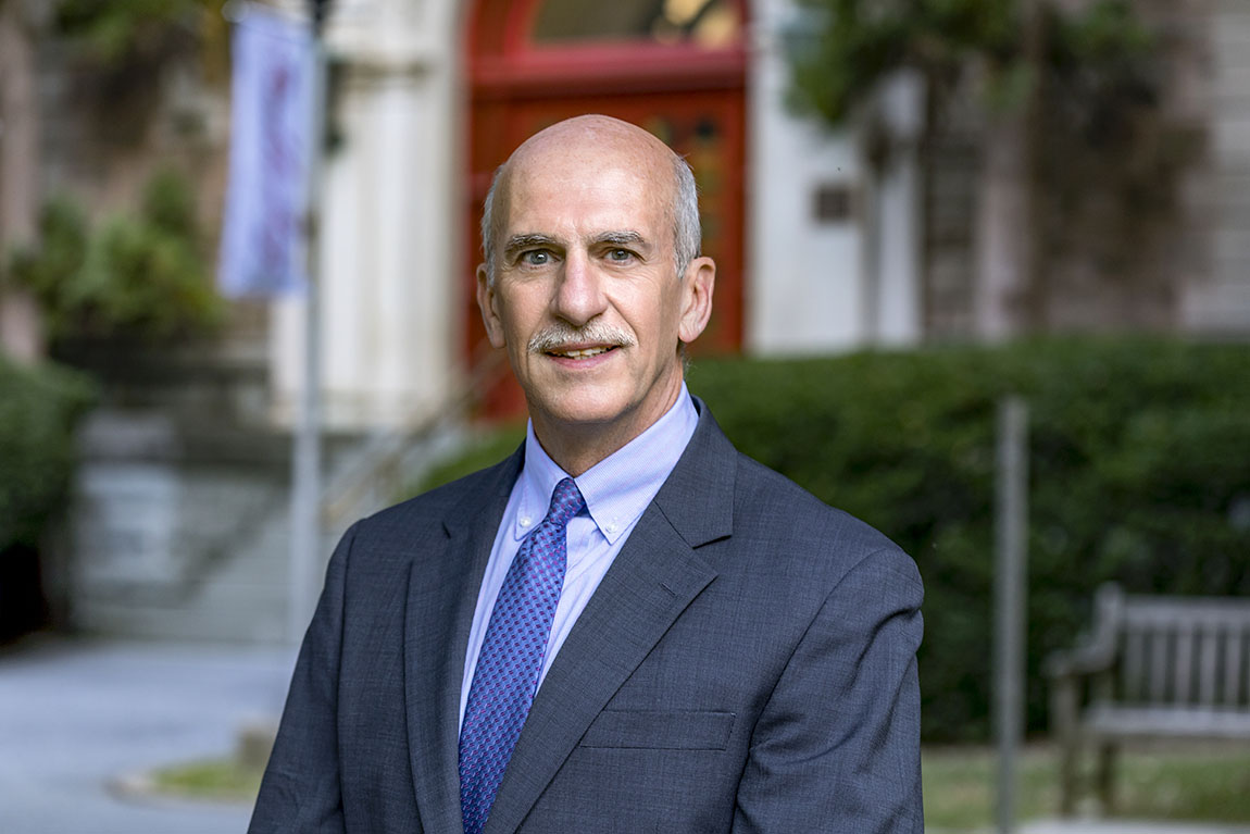 A man with gray hair and a gray mustache wearing a suit jacket and blue tie smiles for a headshot outside on a college campus